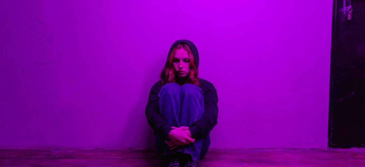 A woman in a hoodie sits alone on the floor, illuminated by purple light indoors.