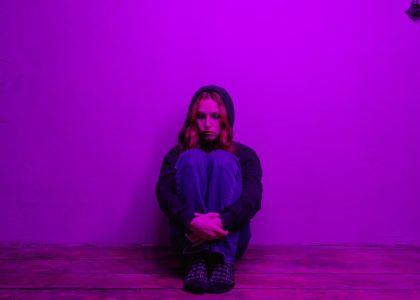 A woman in a hoodie sits alone on the floor, illuminated by purple light indoors.